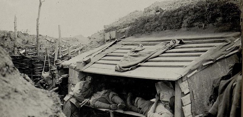 Black and white photograph of 5 Austrian soldiers in a trench, 3 standing, 2 lying in a shelter, 1 of whom is reading a document
