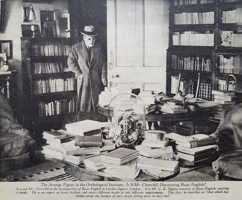 C K Ogden  wearing a white mask that covers his face, a trilby hat and an overcoat. He stands by an open wooden door, in a room, lined with books , behind 2 round tables covered in piles of books.