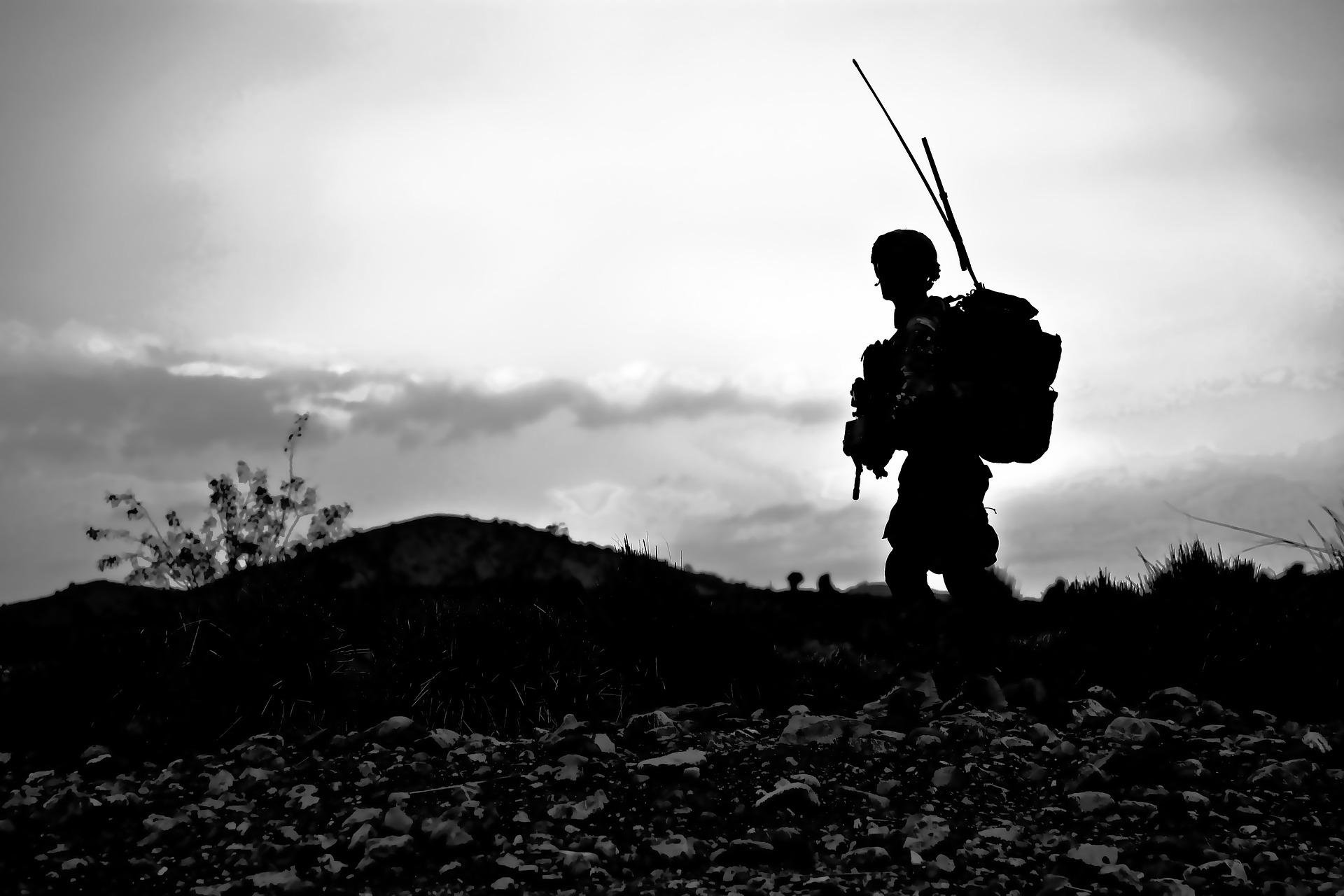 black & white pic of soldier carrying communications equipment with twin aerials silhoutted against bright sky and rocky, hilly landscape with a small scrub. Image by Amber Clay from Pixabay https://pixabay.com/photos/soldier-military-uniform-armed-60762/